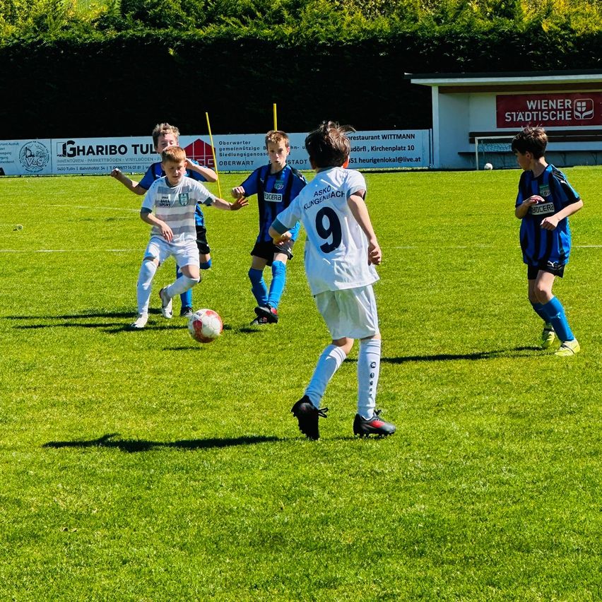 Fünf Jungen in Mannschaftsuniformen spielen Fußball auf einem grünen Feld. Ein Junge jagt dem Ball hinterher, während ein anderer zuschaut. Im Hintergrund befinden sich Torpfosten.