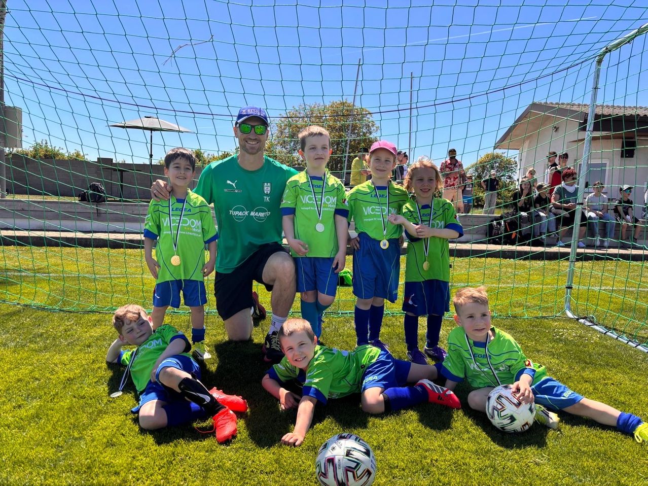 Eine Fußballmannschaft in grünen Uniformen und Medaillen steht vor einem Tor. Ein Trainer in einem grünen Shirt und Hut lächelt. Hinter ihnen ist ein Netz und eine Gruppe von Leuten zu sehen.