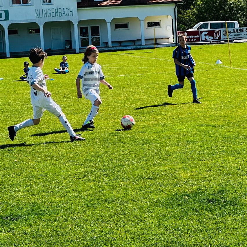 Ein Fußballspiel ist im Gange. Zwei Kinder rennen auf den Ball zu. Ein Kind sitzt hinter ihnen. Ein Gebäude mit dem Wort Klingenbach befindet sich auf dem Feld.