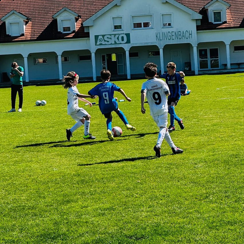 Eine Gruppe von Kindern, die auf einem Feld Fußball spielen, während ein Mann Fotos macht. Sie tragen Fußballuniformen und Turnschuhe. Im Hintergrund befindet sich ein Gebäude mit einem Schild, auf dem 'ASKO KLINGENBACH' steht.