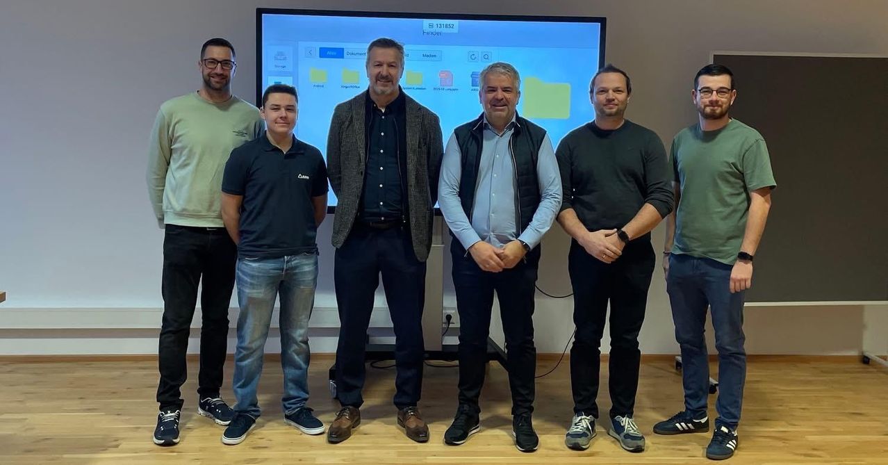 Fünf Männer stehen vor einem Bildschirm, der eine Software-Oberfläche anzeigt. Sie tragen Freizeitkleidung. Der Mann in der Mitte trägt eine Uhr.
