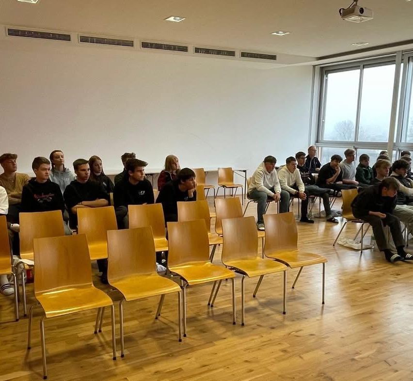 Ein Klassenzimmer mit Holzboden, mehreren gelben Stühlen und einem Beamer. Viele Schüler sitzen in Reihen und schauen nach vorne. Ein Fenster zeigt einen Blick auf einen Baum.