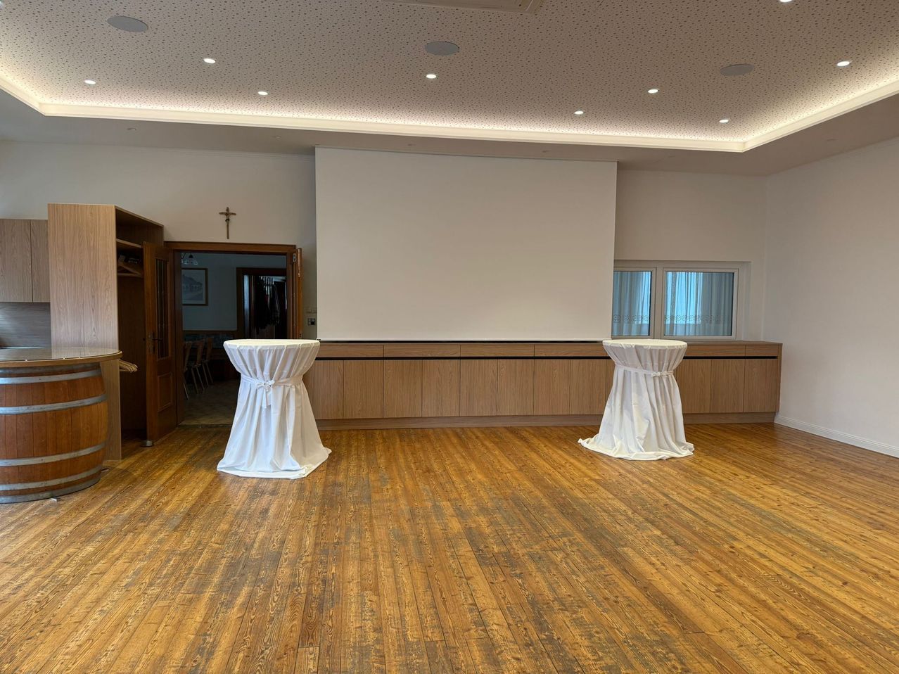 Ein leerer Raum mit Holzböden und zwei in weiße Tücher gehüllte Tische. Eine große weiße Leinwand ist an der Wand montiert.