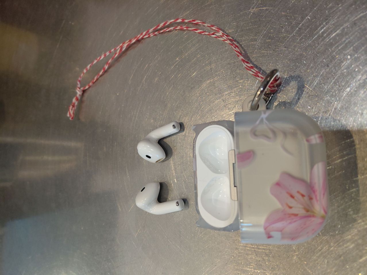 Ein durchsichtiges Wireless-Ohrhörer-Etui mit Blumendesign, geöffnet, um zwei weiße Ohrhörer darin zu zeigen, auf einer metallischen Oberfläche platziert.
