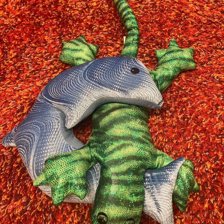 Ein grüner und grauer Plüschgecko liegt auf einem roten Teppich mit seinem Schwanz um seinen Körper gewickelt. Der Gecko hat eine strukturierte, glänzende Oberfläche mit einem fischähnlichen Schwanz.