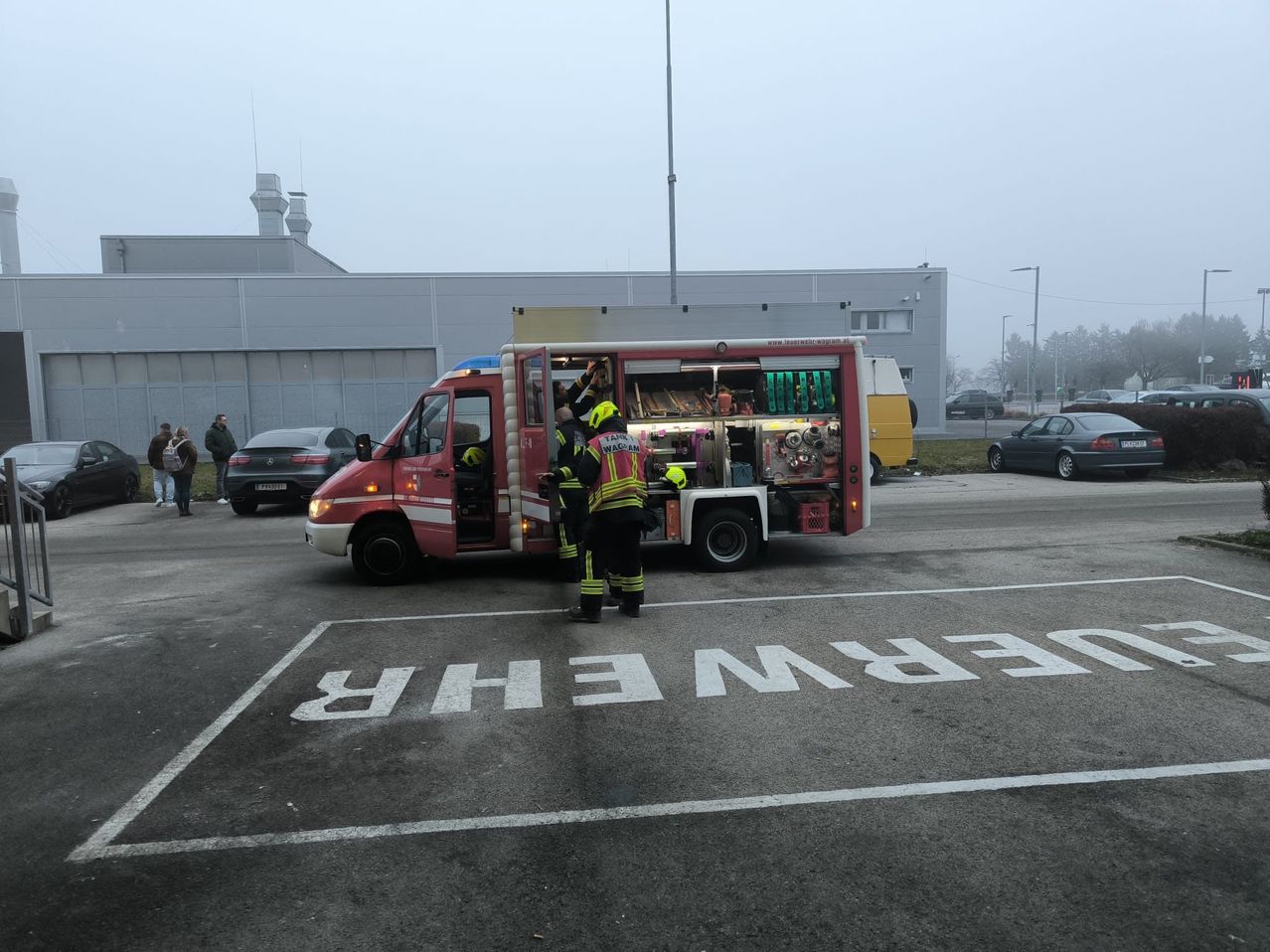 Zwei Feuerwehrleute laden Ausrüstung in ein rotes Notfallfahrzeug, das auf einem Parkplatz geparkt ist. Das Wort 'Notfall' ist auf dem Boden gemalt.