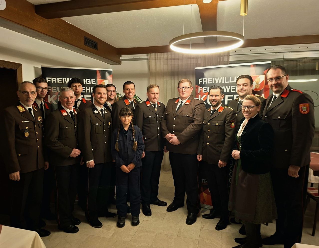 Eine Gruppe uniformierter Personen, darunter Männer und Frauen, posiert für ein Foto mit einem jungen Mädchen vor Bannern mit deutschem Text.