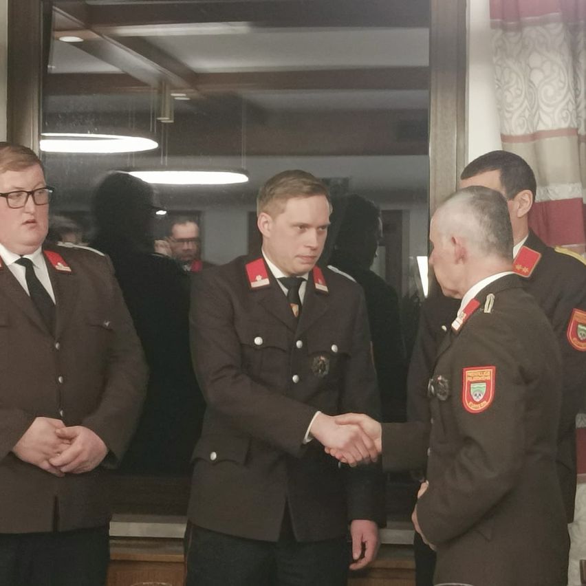 Drei Männer in Uniformen schütteln sich die Hände in einem Raum, wobei zwei weitere hinter ihnen stehen. Ein Spiegel reflektiert einen Raum mit Lichtern und einem Vorhang.