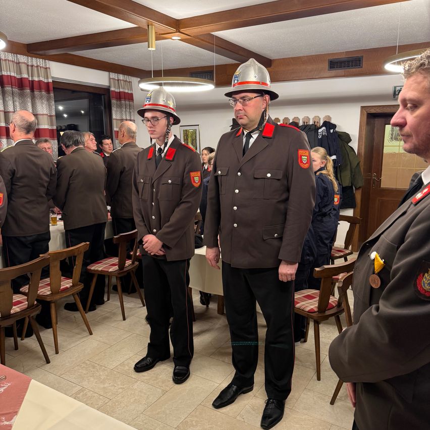Mehrere Feuerwehrleute in Uniform stehen drinnen, einer trägt einen Helm. Sie sind in einem Raum mit gefliesten Böden, Tischen und Stühlen. Ein anderer Mann in Uniform beobachtet sie.