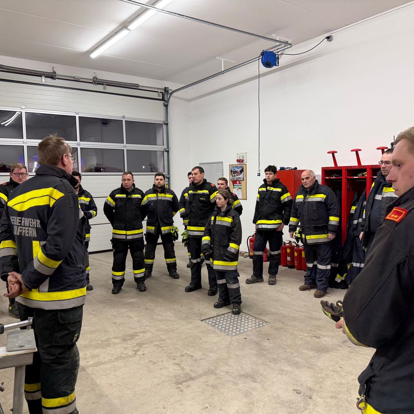 Eine Gruppe von Feuerwehrleuten in Uniformen versammelt sich in einem Raum und hört einer Besprechung zu. Der Raum ist gut beleuchtet mit Lampen an der Decke. Ausrüstung und Feuerlöscher sind an der Seite gelagert.