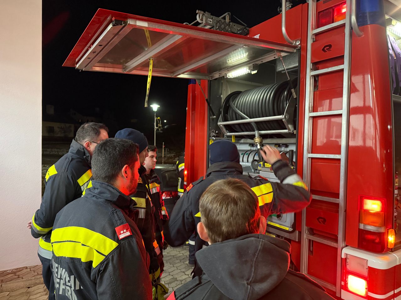 Eine Gruppe von Feuerwehrleuten in Uniform steht bei Nacht um einen Feuerwehrwagen, einer arbeitet drinnen an der Ausrüstung.