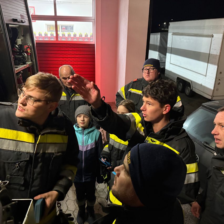 Eine Gruppe von Feuerwehrleuten steht um ein Kind herum und erklärt ihm etwas. Sie sind alle in Uniformen gekleidet und einige tragen Mützen. Ein Feuerwehrauto ist in der Nähe geparkt.