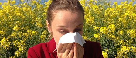 Eine Frau in einem roten Mantel schnäuzt sich in einem Feld voller gelber Blumen an einem sonnigen Tag.