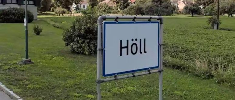 Ein Schild mit der Aufschrift 'Höll' steht am Straßenrand in einem ländlichen Gebiet mit einem Grasfeld. In der Ferne befindet sich ein Gebäude mit einem Turm.