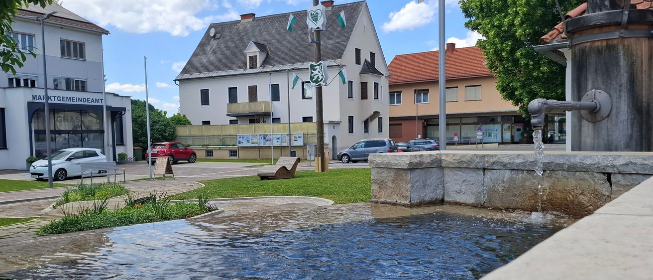 Ein kleiner Stadtplatz mit einem Brunnen, umgeben von Gebäuden und Flaggen. Ein rotes Auto ist auf der Seite geparkt.