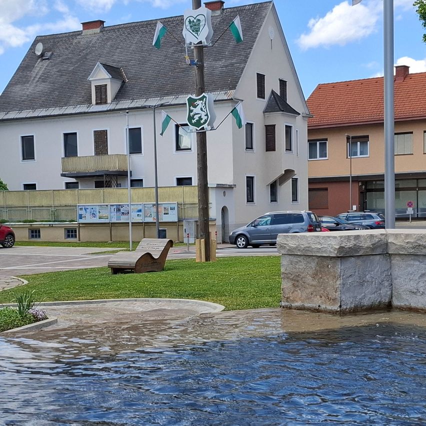 Ein kleiner Stadtplatz mit einem Brunnen, umgeben von Gebäuden und Flaggen. Ein rotes Auto ist auf der Seite geparkt.