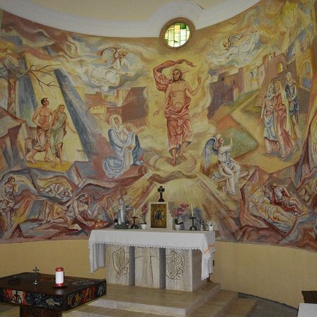 Ein religiöses Wandbild mit verschiedenen biblischen Szenen, darunter Jesus Christus, Engel und Heilige, ist an der Wand einer Kapelle gemalt. Ein Tisch mit Kerzen und Blumen steht vor dem Wandbild.