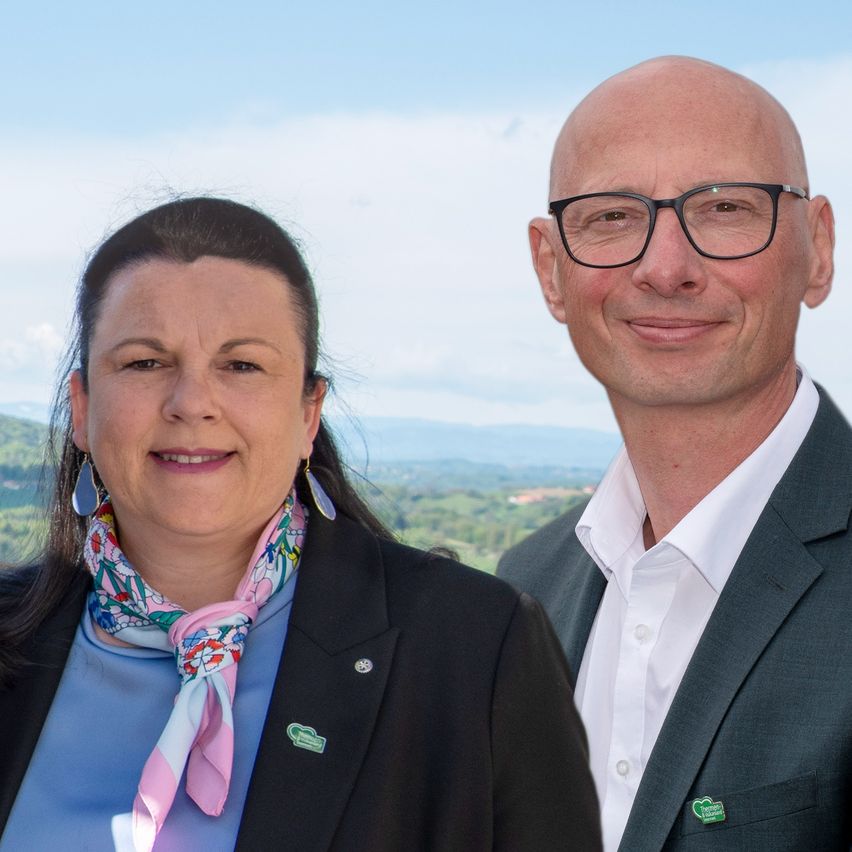Ein Mann und eine Frau stehen lächelnd zusammen vor einer malerischen Landschaft, beide in formeller Kleidung. Die Frau trägt ein Blumentuch und der Mann eine Brille.