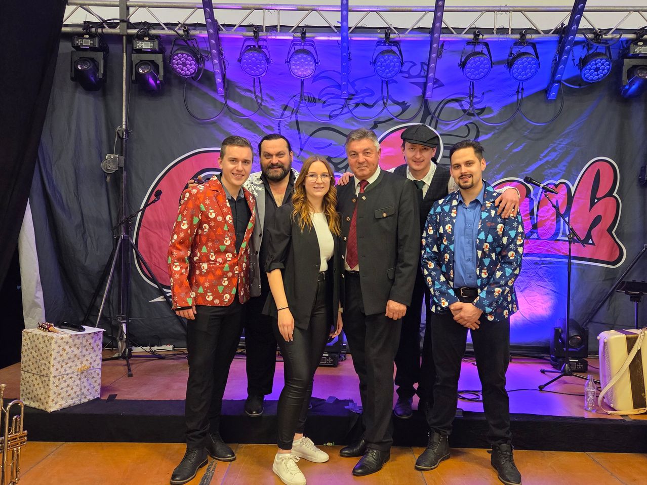 Sechs Personen stehen auf einer Bühne mit einem farbenfrohen Hintergrund. Sie lächeln und scheinen für ein Foto zu posieren. Im Hintergrund befinden sich Scheinwerfer und ein Mikrofon.