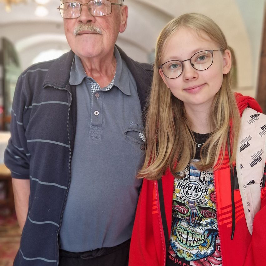 Ein älterer Mann mit einem Schnurrbart und ein junges Mädchen mit Brille und einer roten Jacke stehen eng beieinander und posieren möglicherweise für ein Foto.