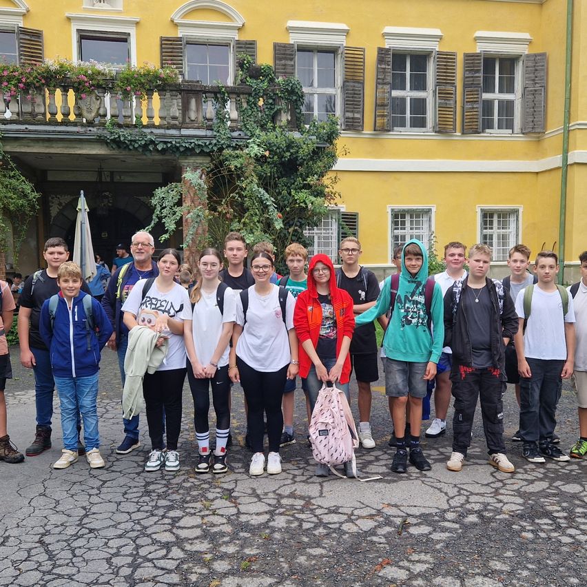 Eine Gruppe von Teenagern und Erwachsenen steht vor einem gelben Gebäude mit vielen Fenstern und einem Balkon mit Pflanzen. Sie sind lässig gekleidet, einige tragen Brillen, und lächeln für ein Foto.
