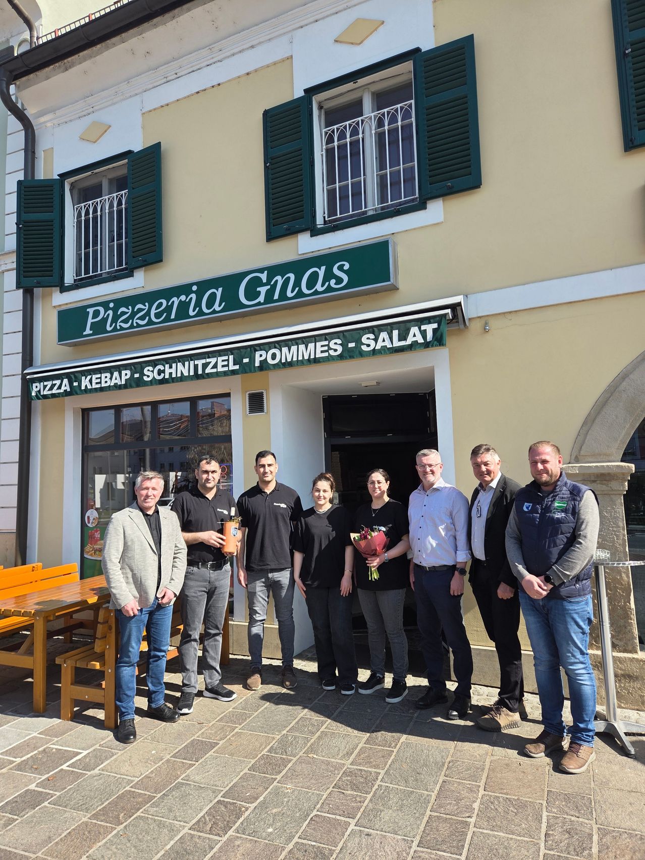 Eine Gruppe von Menschen steht vor einem Restaurant namens Pizzeria Gnas. Sie lächeln und scheinen für ein Foto zu posieren. Links befindet sich eine Holzbank und ein Tisch.
