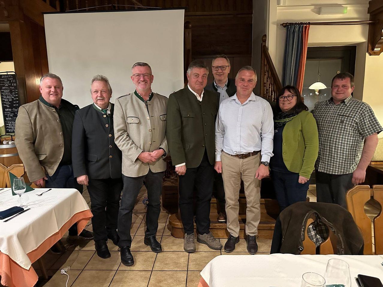 Eine Gruppe von Erwachsenen steht in einem Raum, wahrscheinlich bei einer sozialen Veranstaltung. Sie tragen formelle Kleidung und lächeln für ein Foto. Hinter ihnen befindet sich eine Treppe und ein Tisch mit einer weißen Tischdecke.