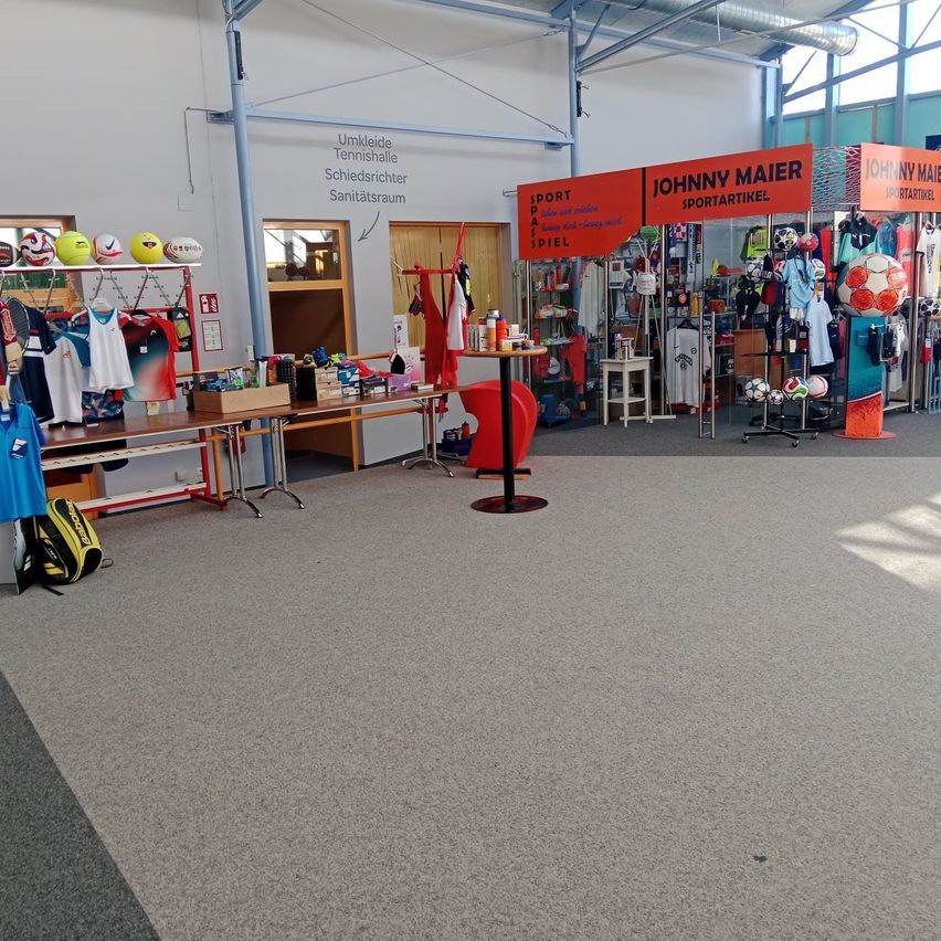Eine Indoor-Sportanlage mit Tischen voller Sportartikel und Ausrüstung, einschließlich Sporttaschen, Bällen und Shirts. Ein Schild an der Wand steht 'Umkleide Tennishalle Schiedsrichter Sanitätsraum'.