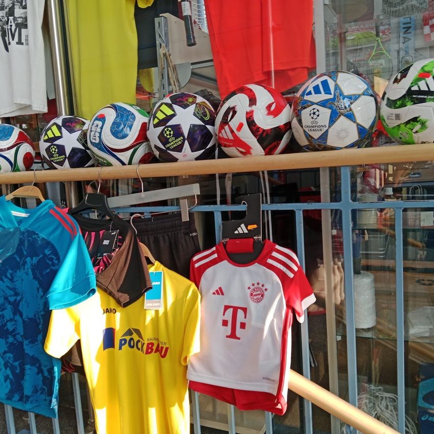 Eine Ausstellung von Fußballbällen, Trikots und Shorts in einem Geschäft. Die Trikots sind für verschiedene Teams, wobei eines die Buchstaben 'T' und ein anderes 'POCI BA' zeigt. Die Fußballbälle sind auf einem Holzregal angeordnet.