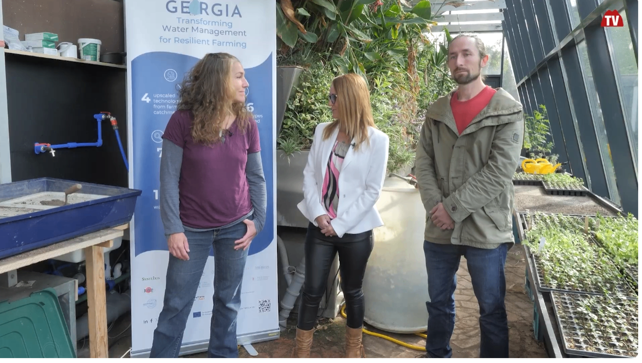 Three people stand in front of a backdrop and a plant. The woman on the left wears a purple shirt, the woman in the middle wears a white jacket, and the man on the right wears a green jacket.