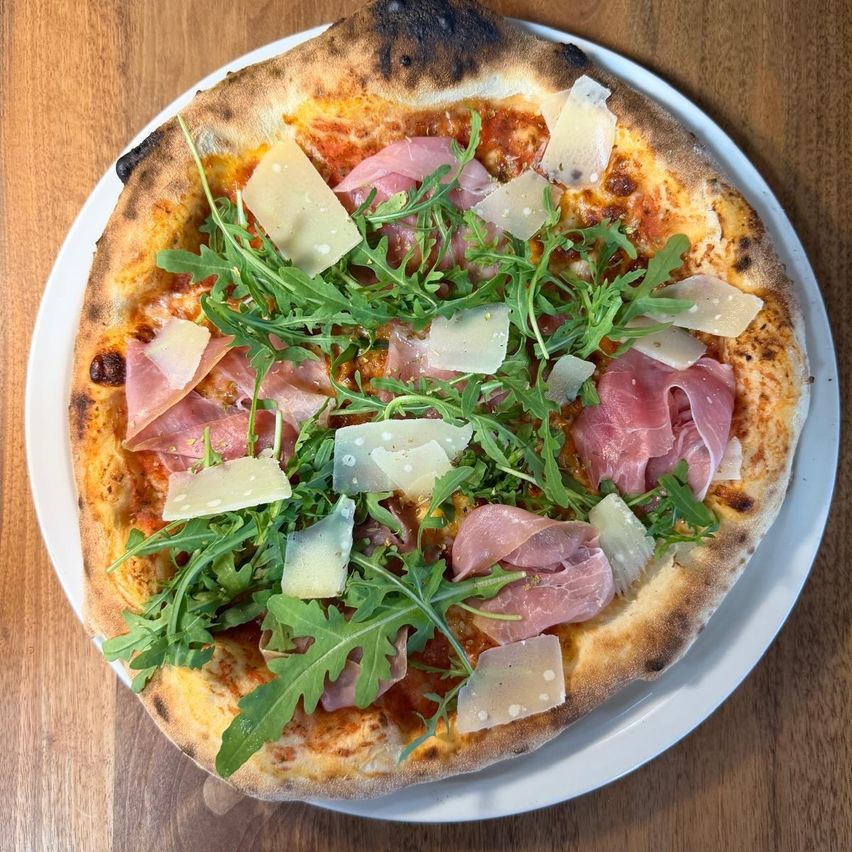 A pizza topped with cheese, arugula, and thinly sliced ham on a white plate on a wooden table.