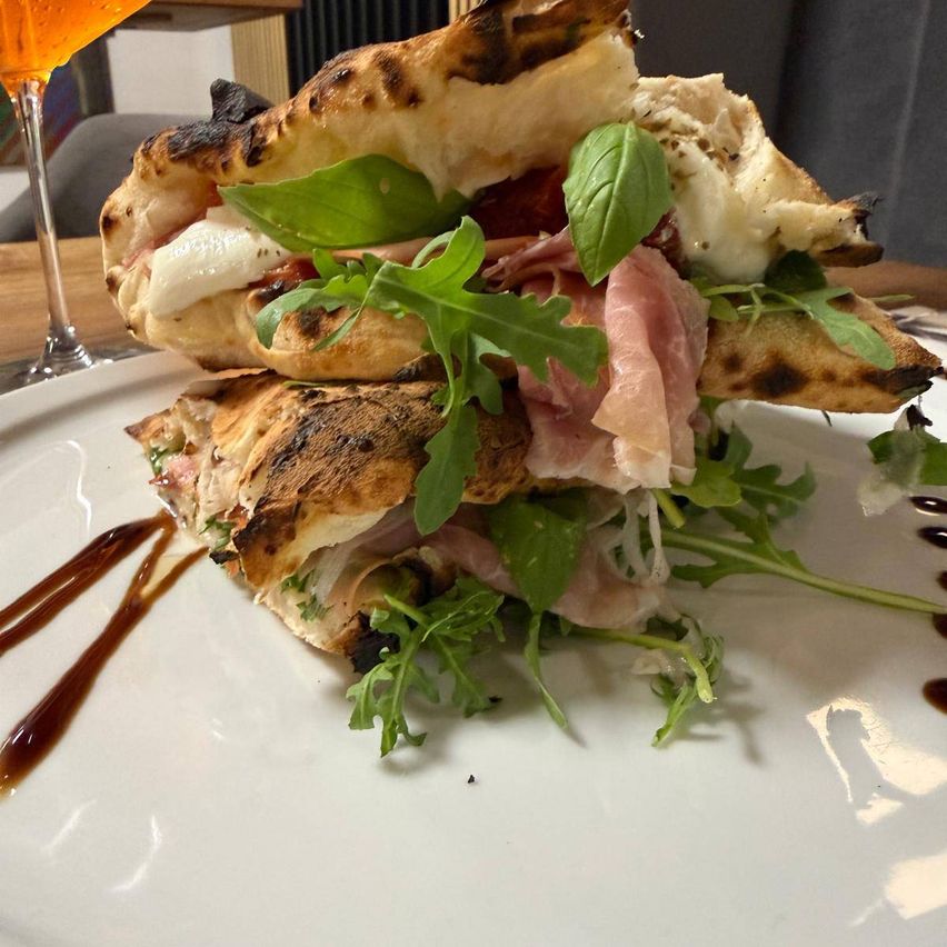 A sandwich with toasted bread, mozzarella, ham, and basil leaves on a white plate with a drizzle of sauce.