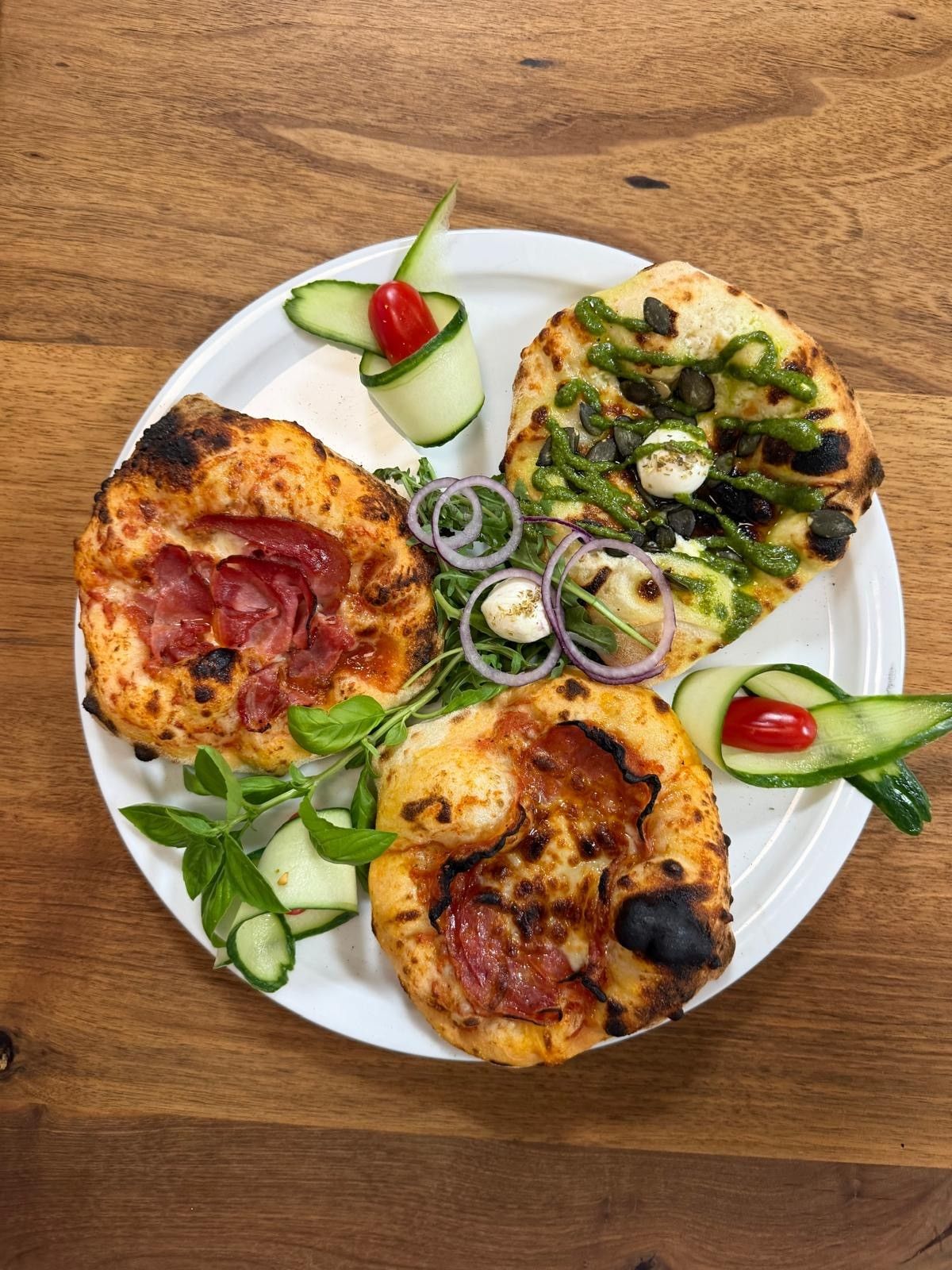 A white plate with three pizzas and a side of vegetables. One pizza has ham, another has green sauce and black olives, and the third has a fried egg.