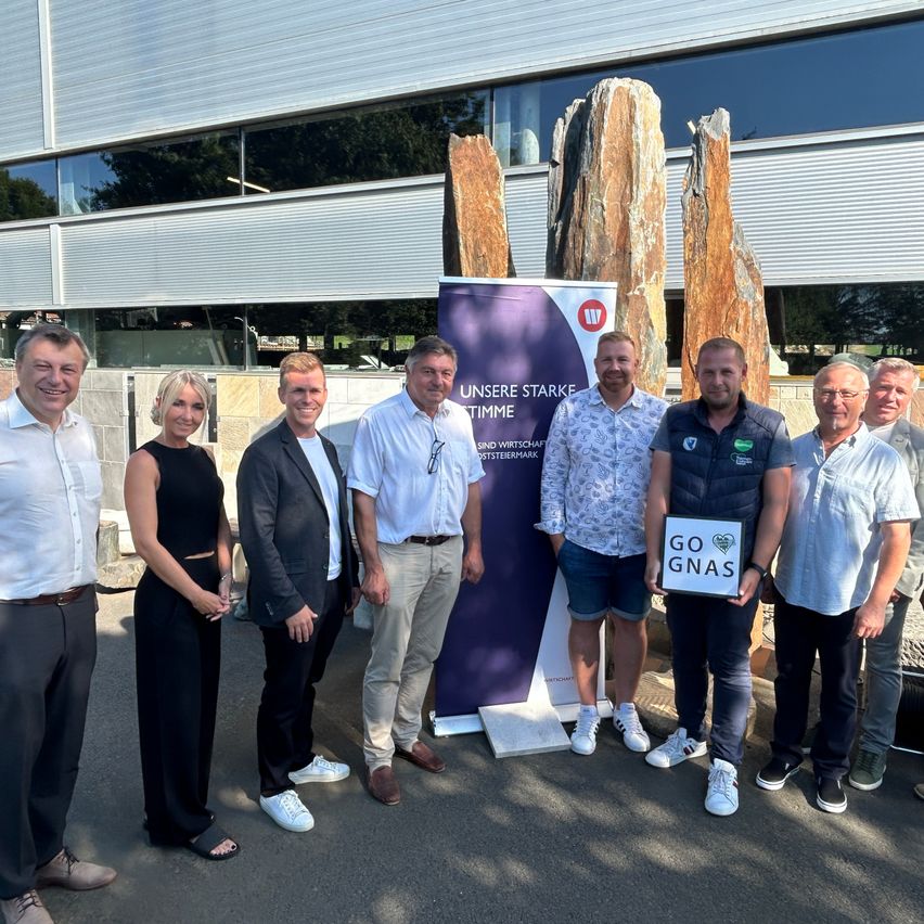 Sieben Personen stehen vor einem Gebäude mit einem Banner, lächeln und posieren für ein Foto. Sie sind in der Nähe von Steinstrukturen.