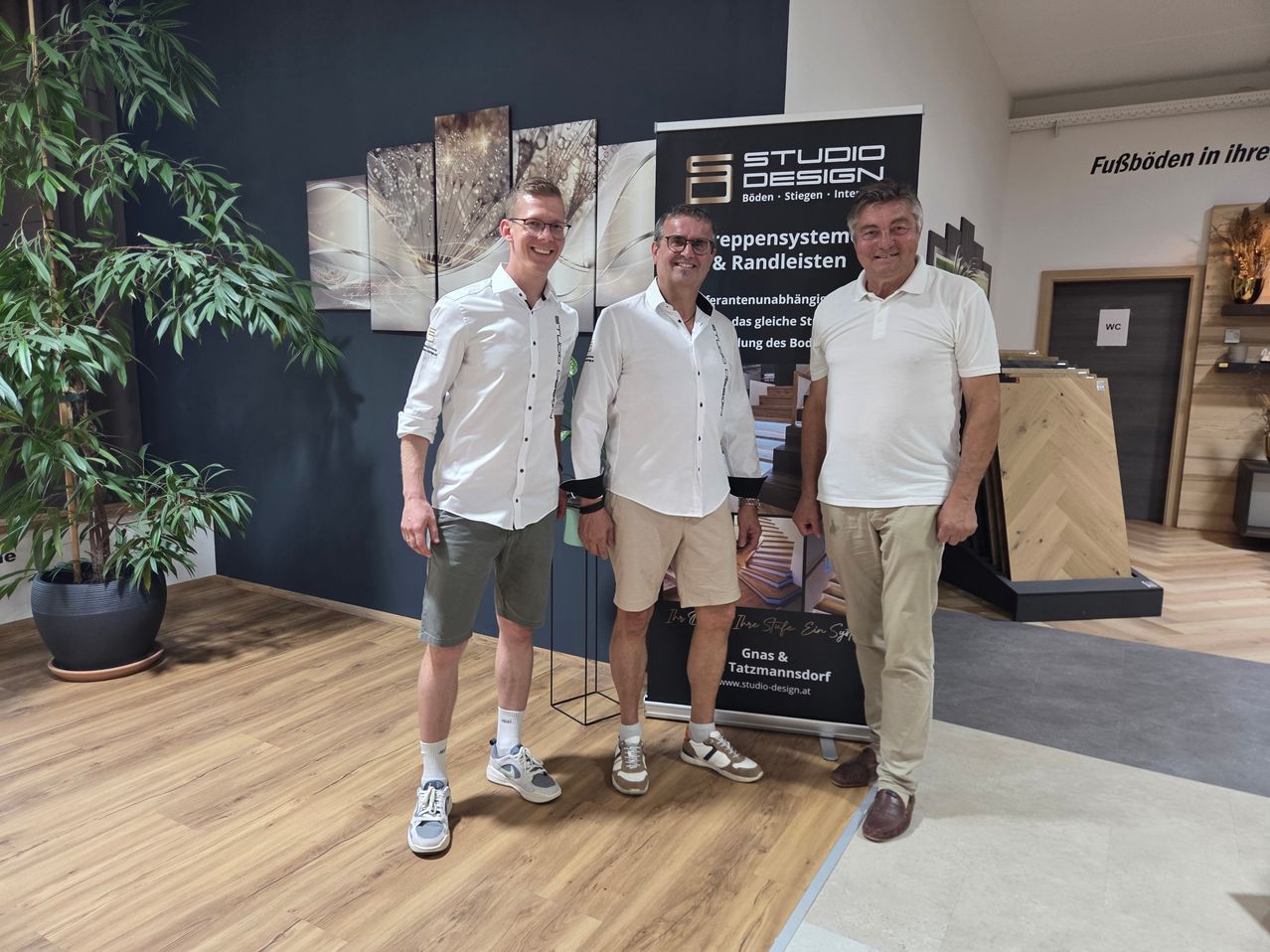Drei Männer in weißen Hemden und Shorts posieren für ein Foto vor einer Wand mit Gemälden und einem Schild.