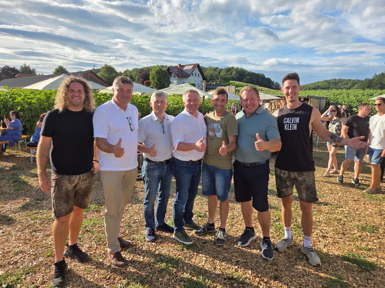Six men pose for a photo on a sunny day in front of a vineyard. They are all smiling and giving thumbs up.