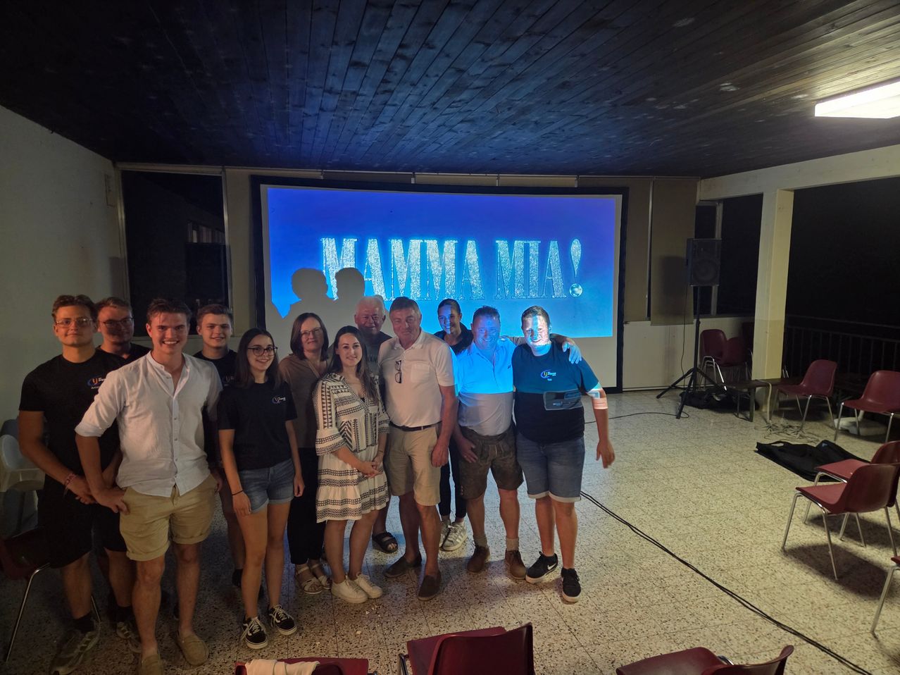 A group of people stands in front of a projector screen displaying 'MAMMA MIA!' with two men in the front wearing blue shirts.