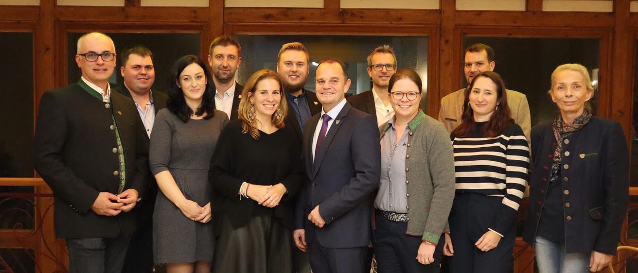 Eine Gruppe von Personen, darunter Männer und Frauen, steht in einem Raum mit einer dekorativen Decke und posiert für ein Foto. Sie tragen formelle Kleidung.