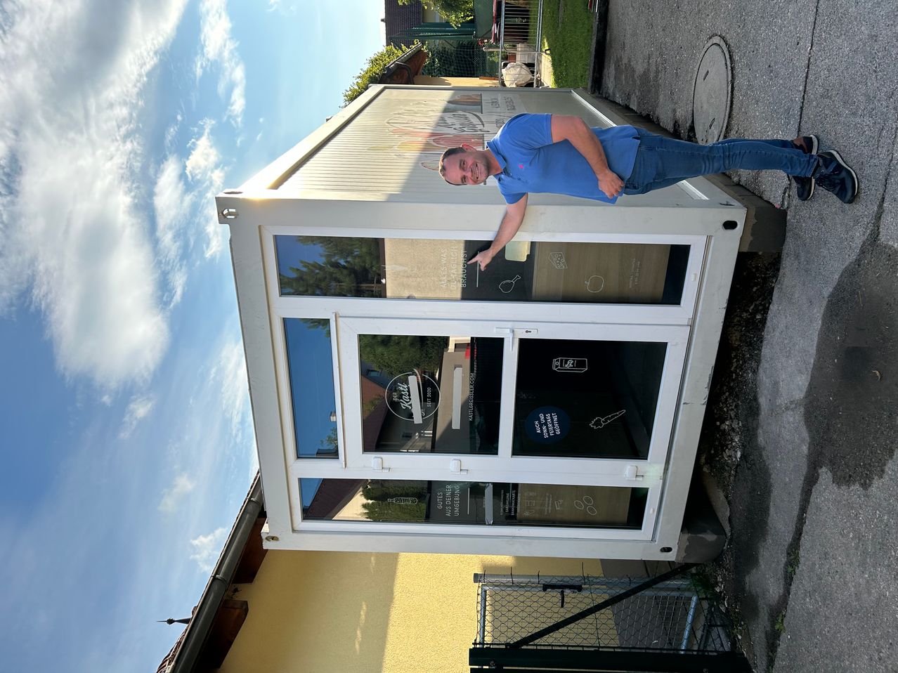 Ein Mann steht vor einem weißen Gebäude mit Glasfenstern und zeigt auf ein Schild, das 'BRAUHAUS' steht. Er trägt ein blaues Hemd und Jeans.
