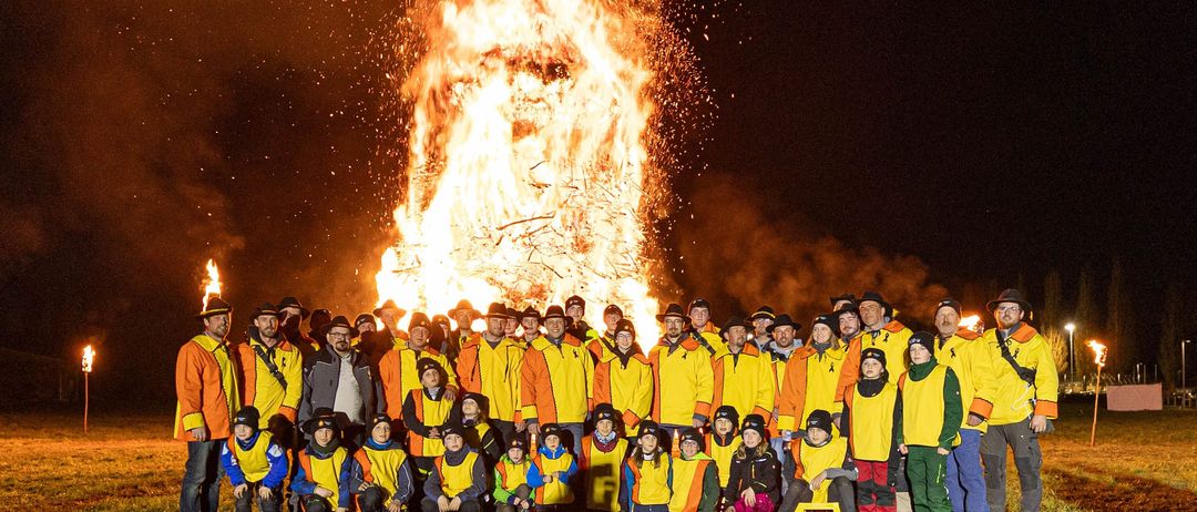 Eine große Gruppe von Menschen in gelben und orangefarbenen Jacken steht vor einem Lagerfeuer bei Nacht.