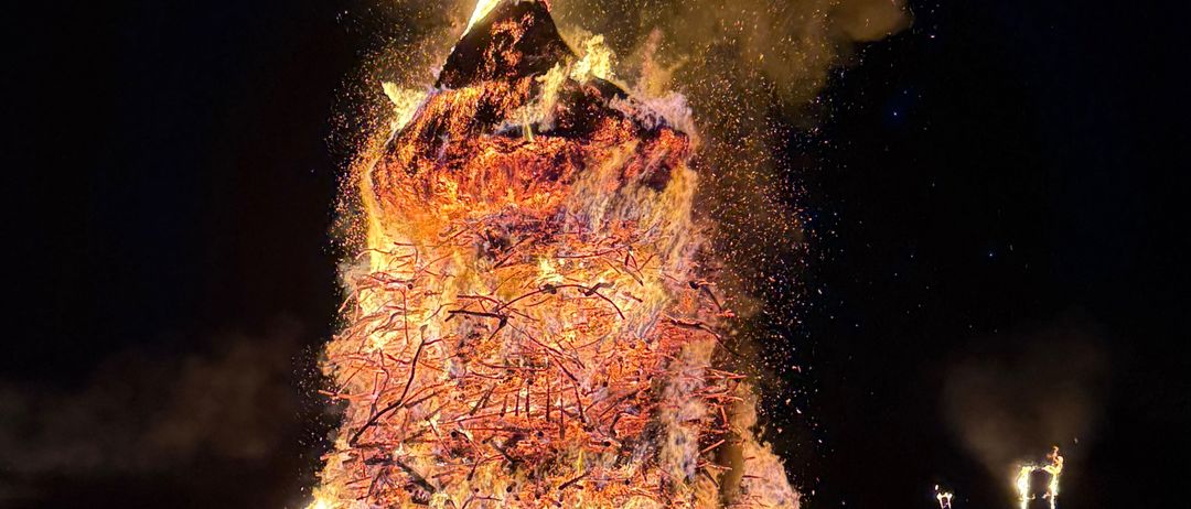 Ein großes Lagerfeuer brennt nachts heftig und wirft einen orangefarbenen Schein und Rauch in den dunklen Himmel. Ein Baumstamm ist auf dem Feuer platziert.