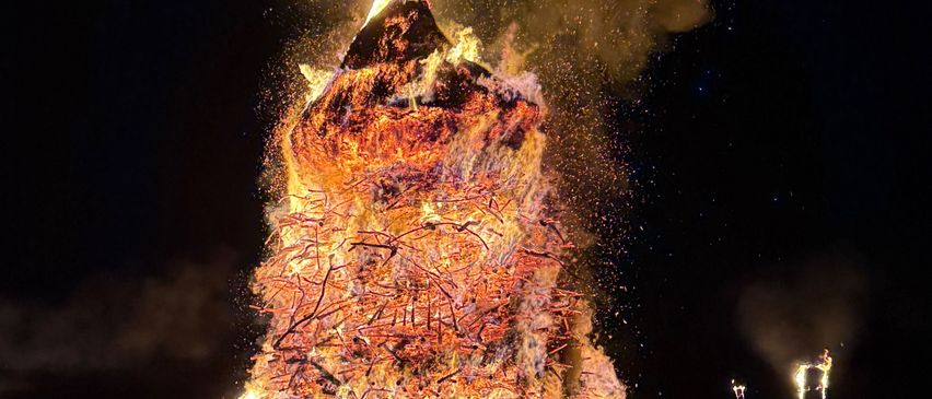 Ein Lagerfeuer brennt bei Nacht hell, mit einem Baum darauf. Helle Flammen und Rauch steigen in den dunklen Himmel. Zwei Laternen erhellen die Szene.