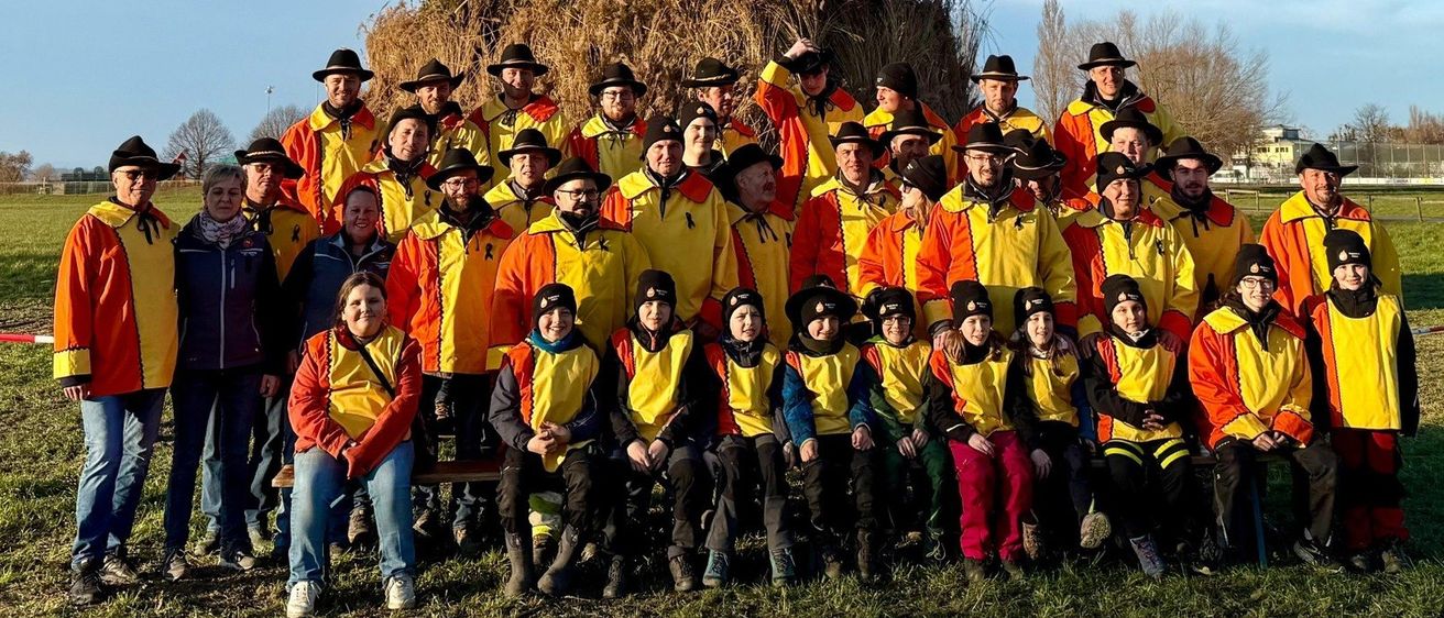 Eine Gruppe von Menschen in leuchtend orangefarbenen und gelben Jacken posiert vor einem großen Strohhaufen mit einem Baum obenauf.