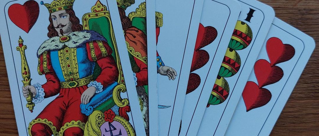 Fünf Spielkarten auf einer hölzernen Oberfläche. Die Karten sind farbig und mit verschiedenen Designs verziert. Die oberste Karte zeigt einen König mit Krone und Zepter.
