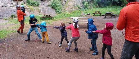 Eine Gruppe von Kindern und Erwachsenen spielt Tauziehen mit einem Seil in einem Waldgebiet mit felsigen Klippen.