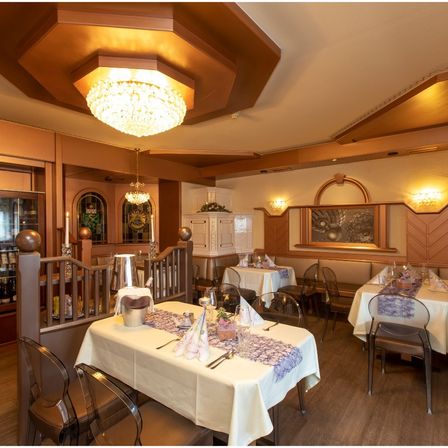 Ein warm beleuchtetes Restaurant mit Holzböden und für das Essen gedeckten Tischen. Es gibt Kronleuchter und an der Wand montierte Lampen, mit einem Gemälde an der Wand.