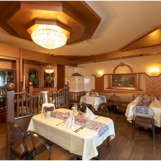 Ein warm beleuchtetes Restaurant mit Holzböden und für das Essen gedeckten Tischen. Es gibt Kronleuchter und an der Wand montierte Lampen, mit einem Gemälde an der Wand.