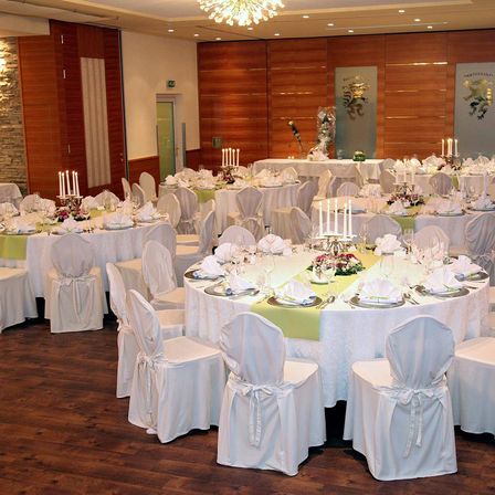 Ein elegant eingerichteter Bankettsaal mit Holzwänden und -böden, der runde Tische mit weißen Tischdecken und verschiedenen Blumenarrangements sowie Stühle, die ordentlich um jeden Tisch herum angeordnet sind, bietet.
