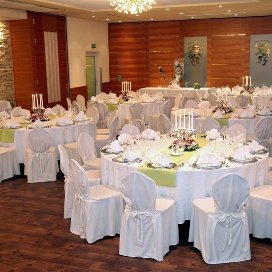 Ein elegant eingerichteter Bankettsaal mit Holzwänden und -böden, der runde Tische mit weißen Tischdecken und verschiedenen Blumenarrangements sowie Stühle, die ordentlich um jeden Tisch herum angeordnet sind, bietet.