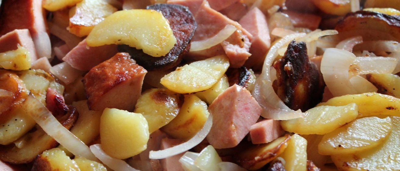 Eine Nahaufnahme eines Gerichts mit gewürfelten Kartoffeln, geschnittenem Schinken und Zwiebeln. Die Kartoffeln sind goldbraun und knusprig, während der Schinken leicht rosa und saftig ist. Die Zwiebeln sind durchscheinend und fügen einen süßen Geschmack hinzu.